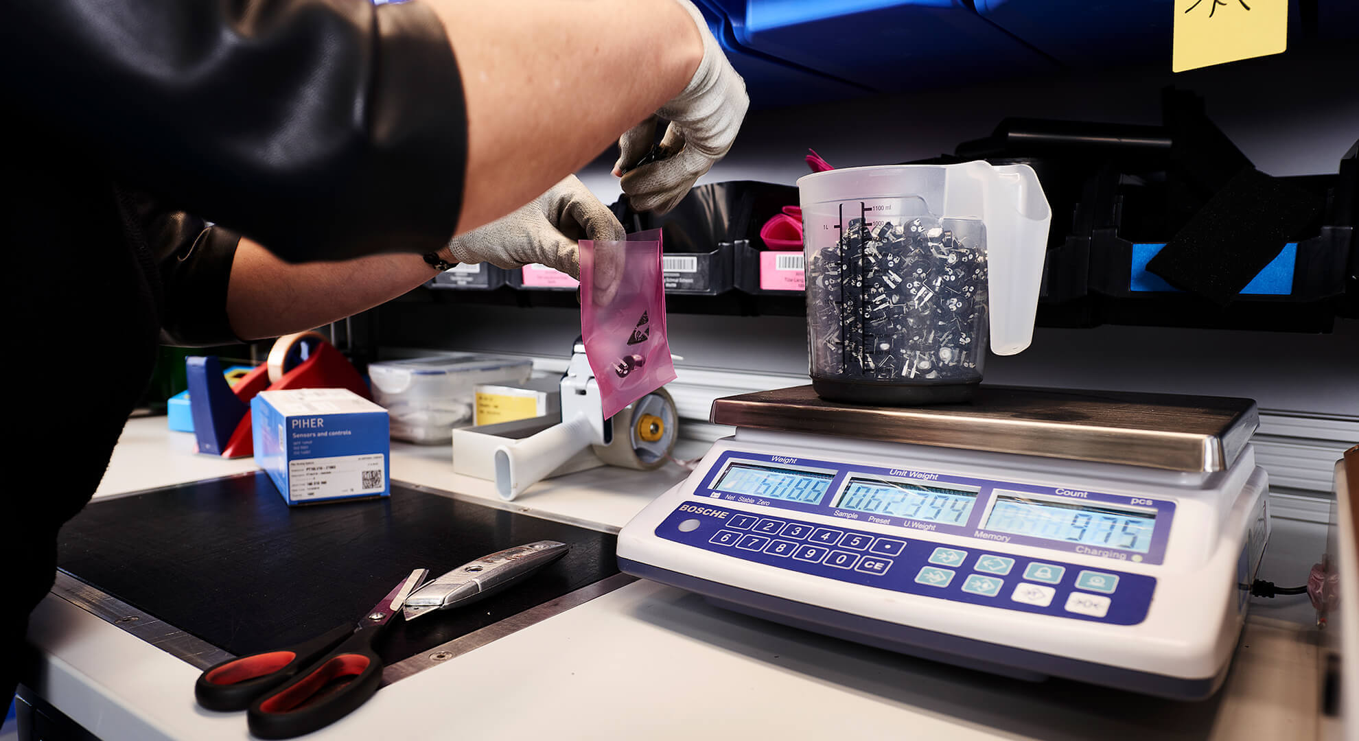 Eine Person mit Handschuhen schüttet kleine schwarze Gegenstände aus einem Messbecher in einen rosafarbenen Beutel auf einer digitalen Waage, während ein OSR Shuttle™ Evo und Verpackungsmaterial, einschließlich einer Schere, in der Nähe auf der Werkbank stehen.