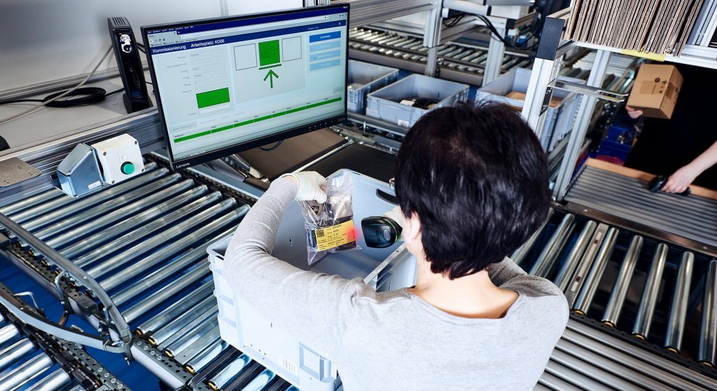 Image from behind of an employee carrying out picking at an ergonomic goods-to-person work station which is equipped with an easyUse interface for intuitive work station design. The woman takes a package of small electronic materials out of a grey storage container. The easyUse user interface shows which articles she needs to remove and where these should be placed. This supports efficient picking processes.