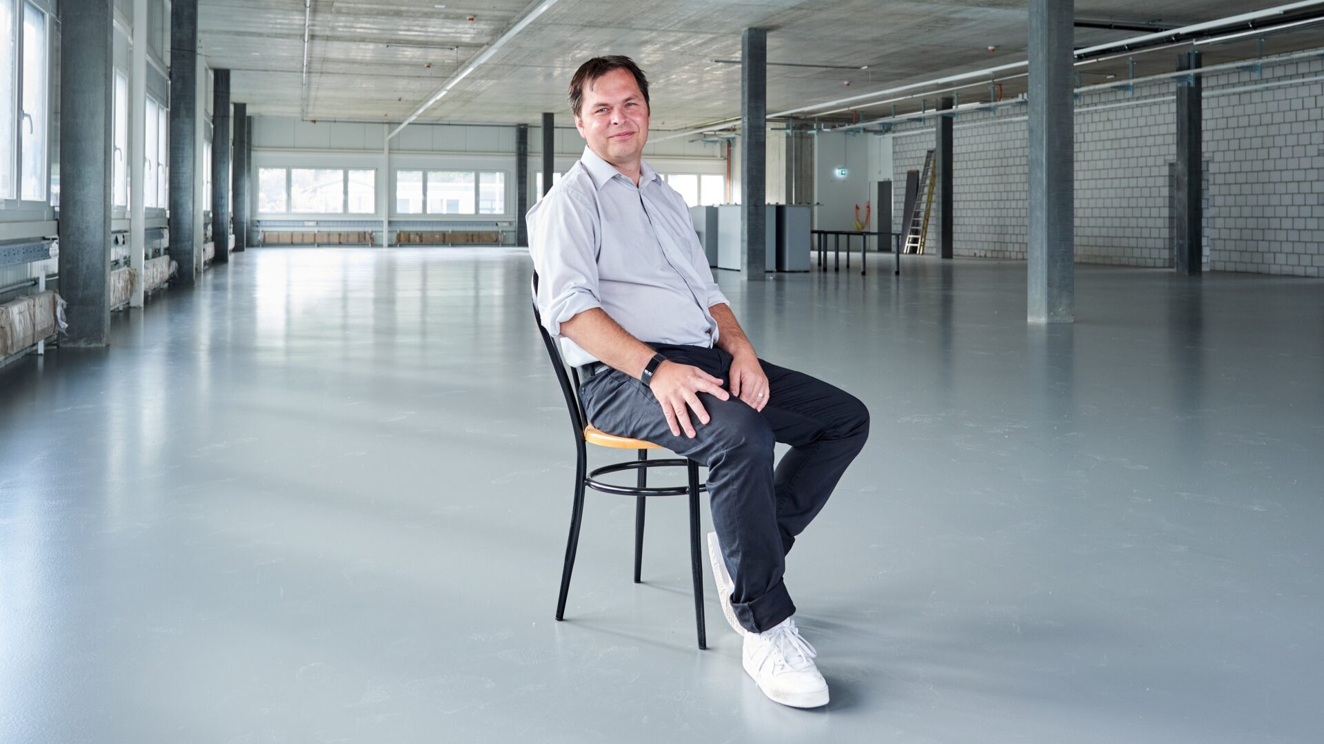 Un homme vêtu d'une chemise claire et d'un pantalon foncé est assis sur une chaise dans une grande pièce vide, de style industriel, dotée de grandes fenêtres.