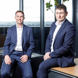 2026 Team Managing Directors Two men in blue suits sit and pose in a modern office discussing warehouse automation for manufacturing, with large windows and a plant behind them.