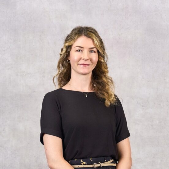 Eine Frau mit welligem blondem Haar, die ein schwarzes Shirt und einen gemusterten Rock trägt und vor einem hellgrauen, strukturierten Hintergrund steht.
