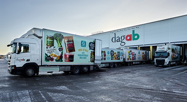 The image shows an outside view of the Dagab Inköp & Logistik AB, Axfood distribution center. In front of the warehouse are several trucks, lining up at the docks to pick up fresh foods for delivery to the stores. The dagab logo is displayed on the building.