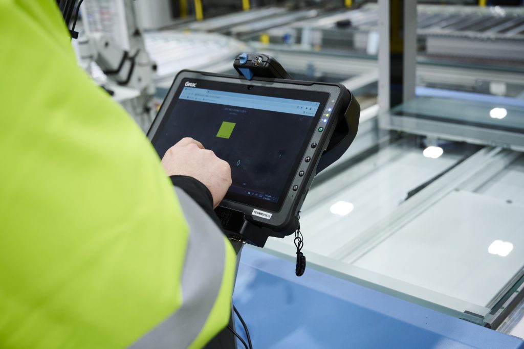 KiSoft Genomix, Appli, Logiciel, Données maîtresses pour l’automatisation, Données maîtresses, Gestion des données maîtresses L’image montre une personne se servant d’une tablette. On voit sur l’écran l’appli KiSoft Genomix. À l’arrière-plan, un carton avec des articles. L’employé saisit au moyen de l’appli toutes les propriétés importantes de l’article qui est représenté à l’arrière-plan.