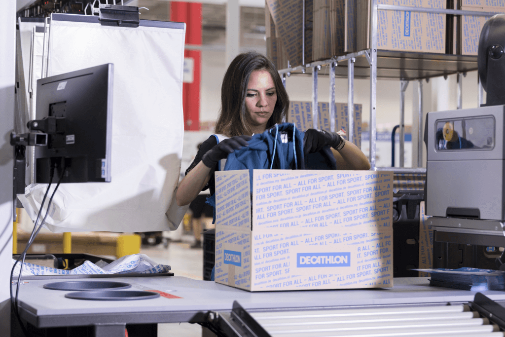 Empleada en el almacén logístico de DECATHLON en una estación de preparación manual con una caja de cartón lista para el envío para pedidos de comercio electrónico.