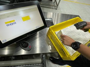 AlternativtextThe image shows a work station in the goods-in area of a pharmaceutical warehouse. Using an RF device, an employee is scanning the serial numbers of medicines, which are in a yellow crate. The SAP® UI5-based interface of the monitor located on the table shows the information on the medicine and the total number of packages that need to be scanned. Besides recording medicines and their serial numbers, SAP® EWM by KNAPP offers further querying functions at these work stations, such as risk-based verification of unqualified partners.