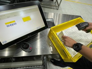 La imagen muestra un puesto de trabajo en el área de entrada de mercancía en un almacén farmacéutico. Un empleado escanea con un radioterminal los números de series de medicamentos que se encuentran en una bandeja amarilla. En la pantalla al lado se indica en una interfaz basada en SAP® UI5 la información correspondiente al medicamento, así como la cantidad de unidades de este medicamento que debe registrarse. SAP® EWM by KNAPP contiene en estos puestos de trabajo, además del registro de medicamentos y sus números de serie, posibilidades de consulta adicionales como la verificación en función de los riesgos de un socio no cualificado.