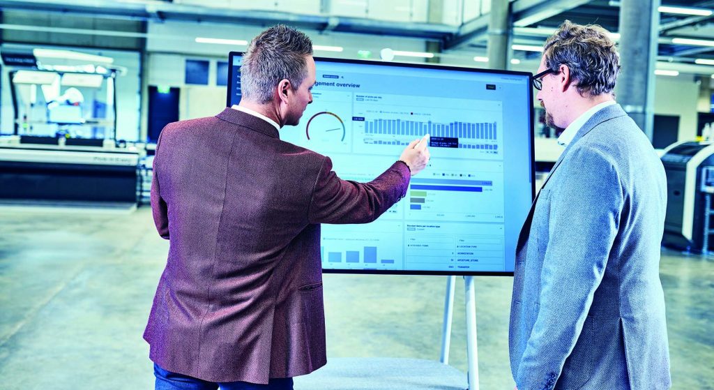 Two men are standing in front of a screen and discussing the displayed charts.