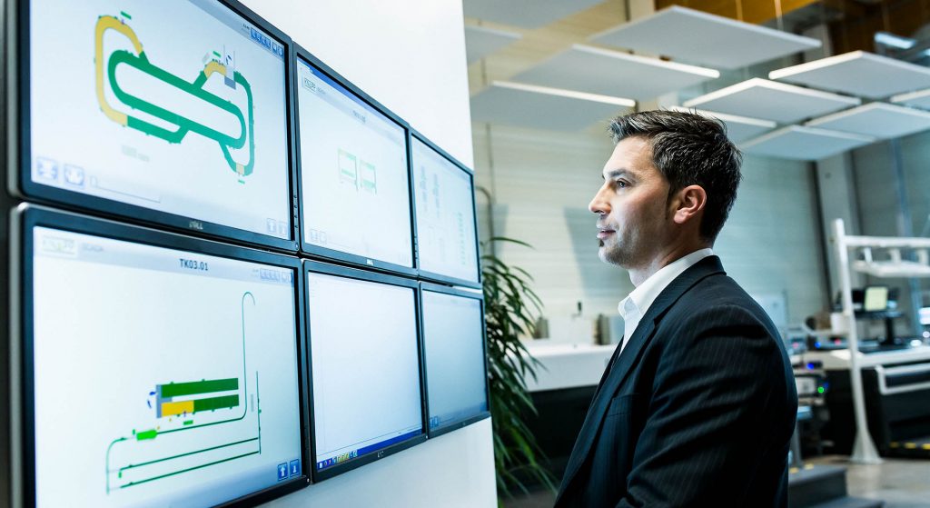 A man is standing in front of a wall of screens showing the WMS and WCS with information on components