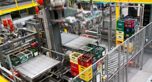 The image is an overview of our sorting system at EDEKA Rhein-Ruhr/t-log. Stacked beverage crates can be seen in the foreground. A robot depalletizes beverage crates layer by layer and transfers them to a conveyor for further processing.