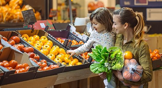 The image shows a young mother with a ponytail holding her four year old daughter on her hip. The two are in a supermarket and buying fresh fruits and vegetables. The mother is holding lettuce and yellow tomatoes in her left hand. With her right hand, the child is reaching into a crate that is filled with yellow tomatoes. Both are smiling happily. To the right and left of the yellow tomatoes are crates holding red tomatoes.