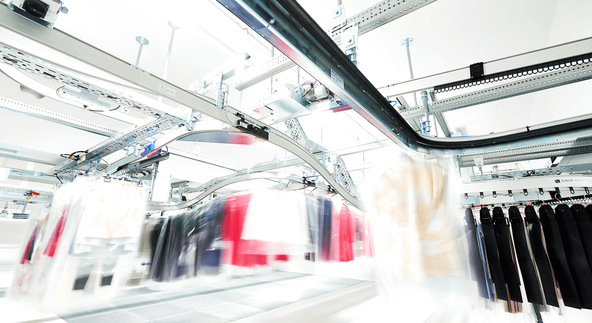 Punto de convergencia de mercancías del sistema de transporte suspendido en Marc Cain Se muestra una nave con un sistema de transporte suspendido de Dürkopp Fördertechnik en movimiento con prendas coloridas. El sistema de transporte suspendido se encuentra en el centro de distribución para moda de Marc Cain.