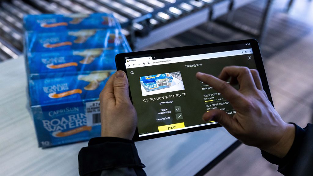 La imagen muestra a una persona usando una tablet. En la pantalla se ve la aplicación KiSoft Genomix. En segundo plano, una caja de cartón con artículos. El operario registra con ayuda de la aplicación todos los atributos relevantes del artículo reflejado en segundo plano.