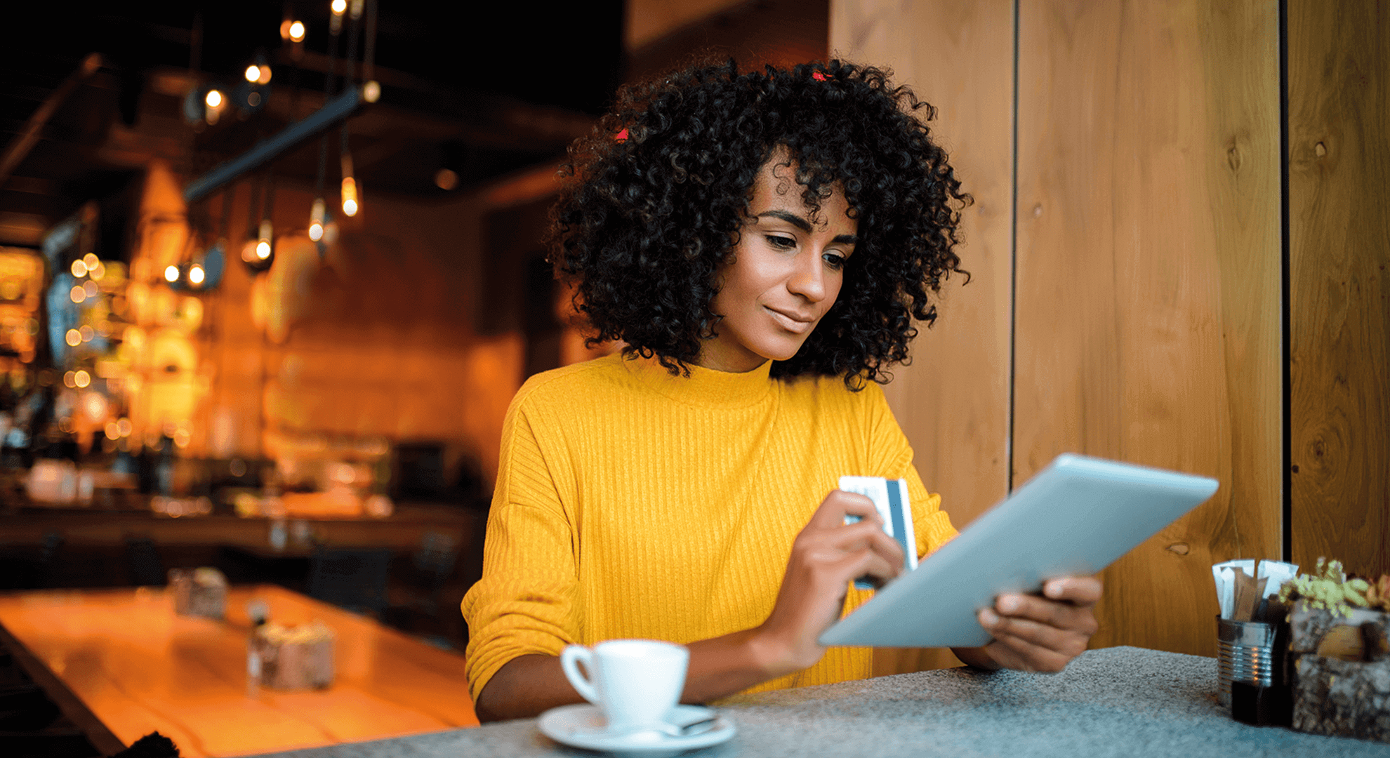 A woman is sitting at a bar with a tablet, drinking a cup of coffee while shopping online. So that her order will arrive quickly, intelligent e-com fulfillment and automation for e-commerce is required