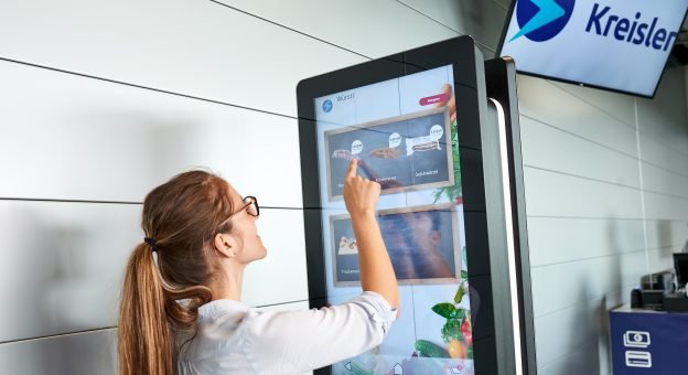L’image montre une jeune femme vue de dos qui choisit les aliments qu’elle souhaite acheter sur un grand écran au moyen d’un écran tactile puis qui les met dans un panier virtuel.