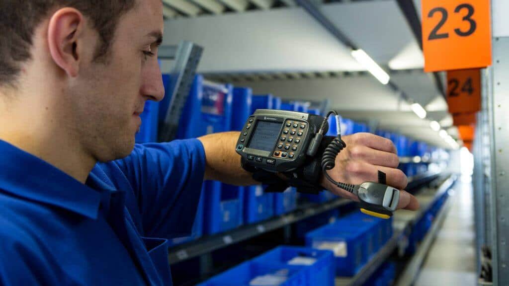 Un collaborateur regarde un terminal radio pour le prélèvement RF accroché à son bras. Le terminal de prélèvement RF aide à la préparation de commandes sans utilisation de papier et indique à l’opérateur où les différents articles se trouvent.