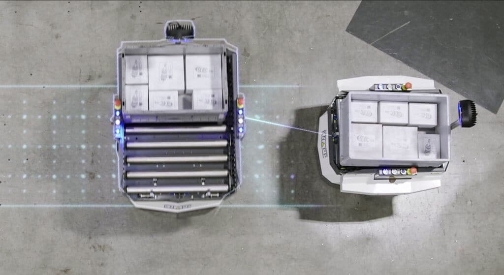 Zwei autonome mobile Roboter, die Open Shuttles begegnen sich in einer Lagerumgebung. Die intelligenten Transportroboter (AMR) sind mit Sensoren ausgestattet und können sich so sicher ausweichen. Jedes Open Shuttle transportiert gerade einen Behälter mit Waren. Autonome mobile Roboter übernehmen zuverlässig innerbetriebliche Transporte.