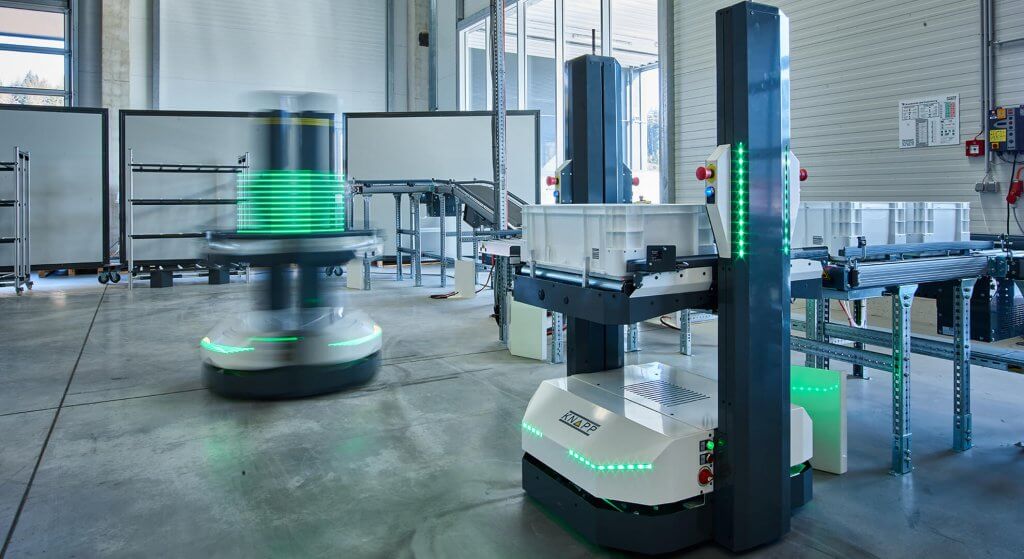 Open Shuttles – autonome mobile Roboter Zwei Open Shuttles transportieren unterschiedliche große Behälter in einem Lager. Open Shuttles sind autonome mobile Roboter (AMR), die für verschiedene Aufgaben in Distributionslägern und Produktionen eingesetzt werden können. Die AMR benötigen keine Leitlinien oder Landmarken und finden ihren Weg im Lager selbstständig.