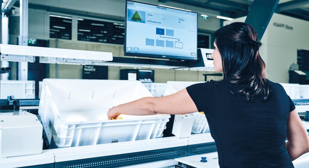 Woman during picking standing in front of an ergonomic goods-to-person work station, the Pick-it-Easy work station from KNAPP, and picking goods from a container. The goods are delivered to the woman using the goods-to-persons (GtP) principle. She is looking at a touch screen that is equipped with the easyUse user interface. This makes work at the work stations easier and optimizes quality and usability. The Pick-it-Easy picking work station improves ergonomics for employees in the warehouse.