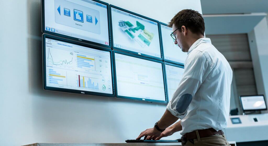 CMMS and SCADA Man standing in front of several screens and using the KiSoft SCADA software with the mouse and keyboard. This is connected to the redPILOT Operational Excellence MAINTENANCE CMMS via an interface.