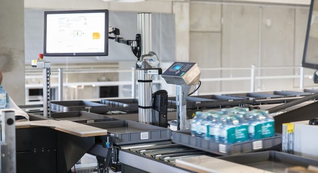 The image shows an ergonomic Pick-it-Easy Case work station with a monitor. The monitor is a visual aid to the employees as they assemble orders. The image also shows a tray with mineral water.