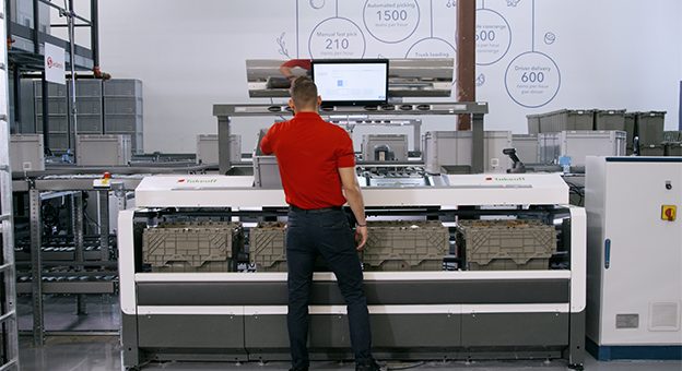 The image shows a Pick-it-Easy work station and an employee with his back to the camera. The employee prepares e-commerce orders for shipping at this ergonomic work station. A screen shows all the steps that he must carry out in this process. He receives precise instructions, for example, which item should be positioned in which bag. After completion, the grocery bags are ready for delivery to the consumer.