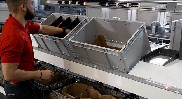 Comercio minorista de comestibles, comercio en línea de comestibles, comercio electrónico de comestibles, Pick-it-Easy workstation, puesto de trabajo Pick-it-Easy, E-Grocer MFC, centro de micrologística, Micro Fulfillment Center, picking, preparación La imagen muestra una vista detallada de nuestros puestos de trabajo Pick-it-Easy con un operario de lado. Mira a una pantalla que indica qué alimentos debe colocar en qué bolsa. Con su mano izquierda, coloca un artículo en una caja.