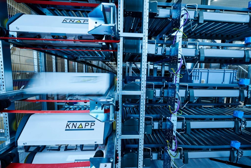 View into the rack line system of the automatic storage system, the OSR Shuttle™. Shuttles are retrieving containers. A shuttle has wheels permitting lengthwise and crosswise movements in the rack. An LED bar is located at the front of the shuttle. The OSR Shuttle™ can be used in different industries.