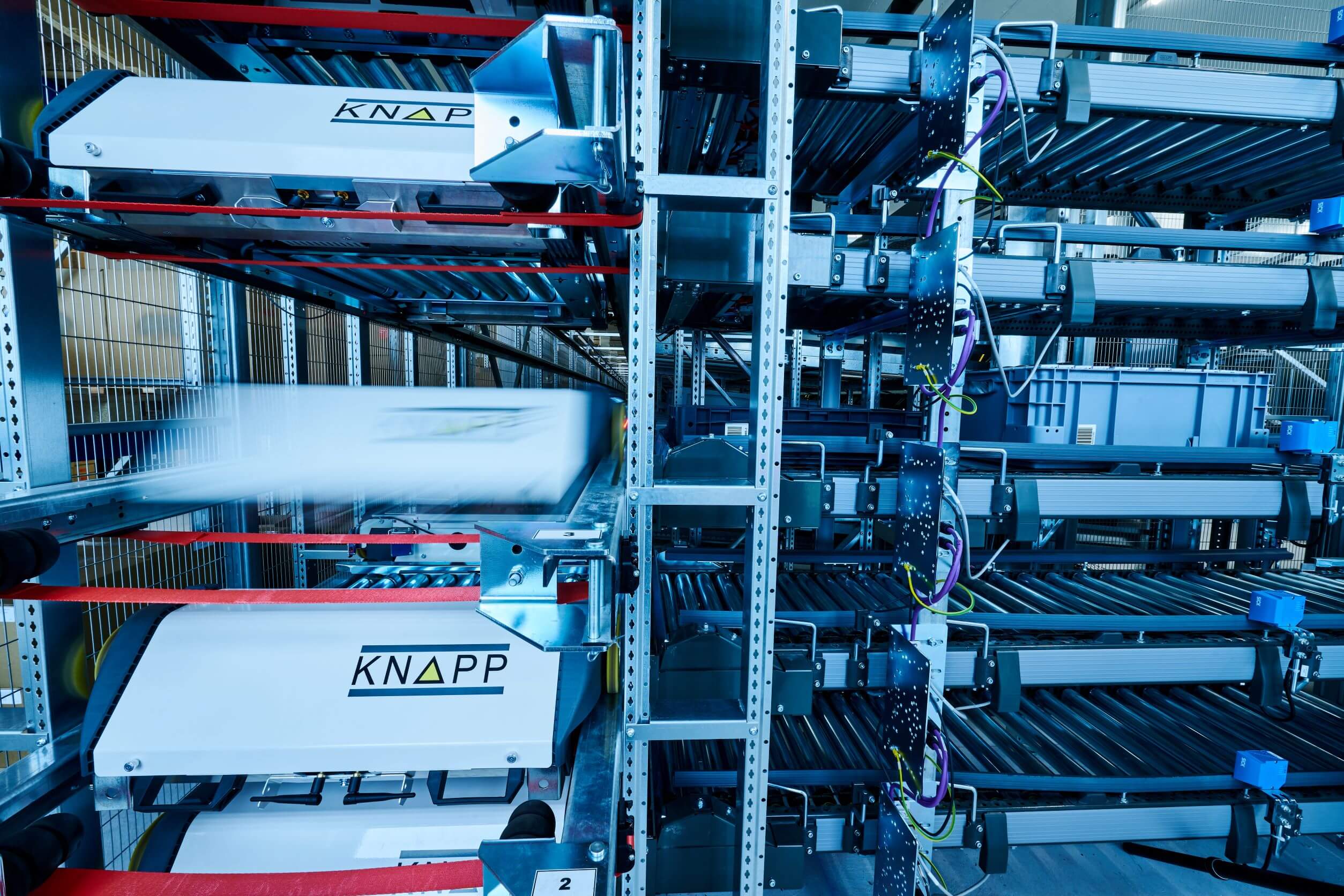 Vista del sistema de líneas de estanterías de un sistema de almacenamiento automático OSR Shuttle™. Las lanzaderas están desalmacenando cajas. La lanzadera tiene ruedas con las que puede desplazarse de manera longitudinal y transversal. En la parte delantera de la lanzadera hay una tira de ledes. El OSR Shuttle™ se puede aprovechar en diferentes sectores.