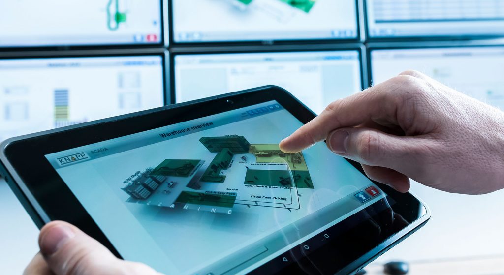 A person is using the touch display on a tablet, which shows the components of a warehouse system