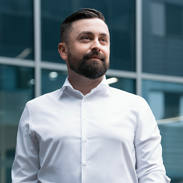 Ein bärtiger Mann mit kurzen dunklen Haaren und weißem Hemd steht selbstbewusst vor einem modernen Glasgebäude.