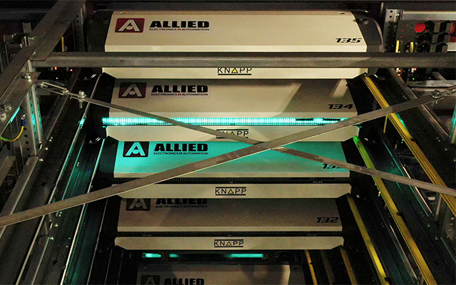Four automated storage and retrieval system shuttles, labeled “ALLIED” and “KNAPP,” are stacked in a warehouse rack, illuminated by green lighting and metal railings.
