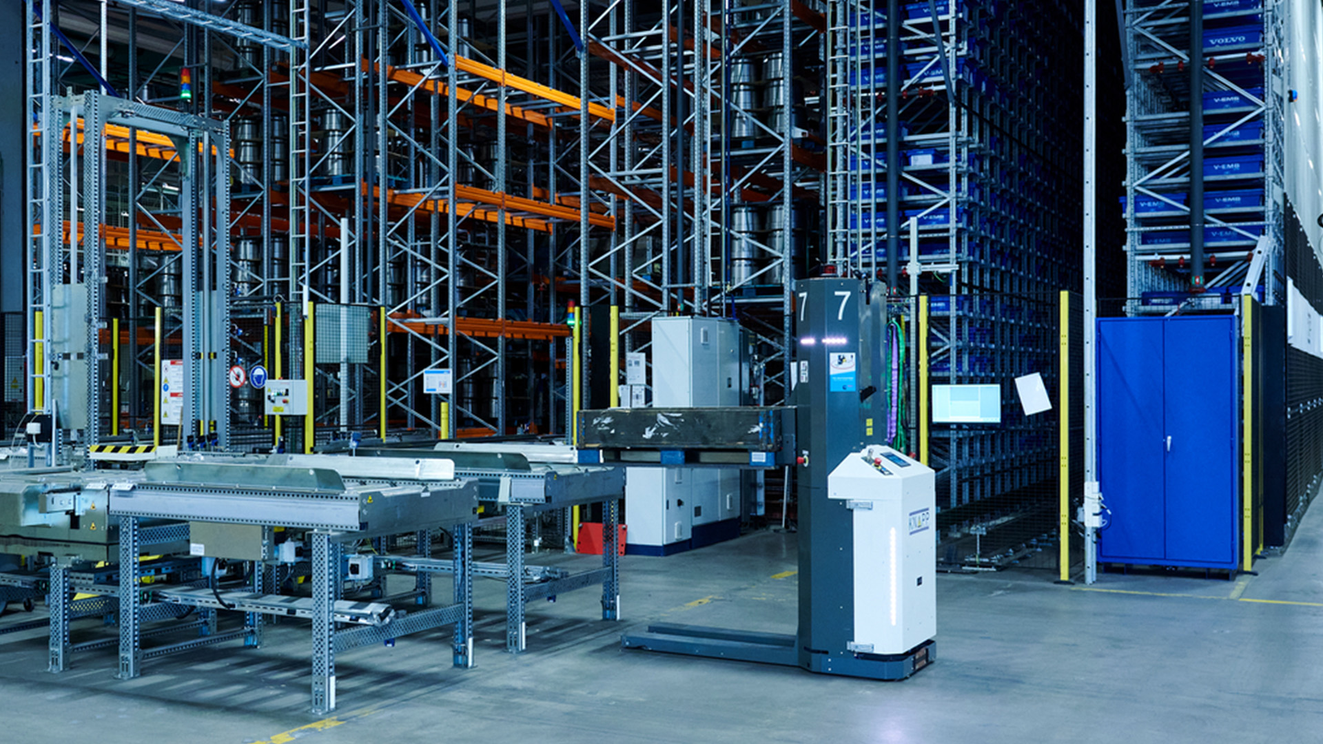 AMRs connect to pallet storage systems A warehouse with tall storage racks, a conveyor system, and an automated guided vehicle moving materials on a clean concrete floor.