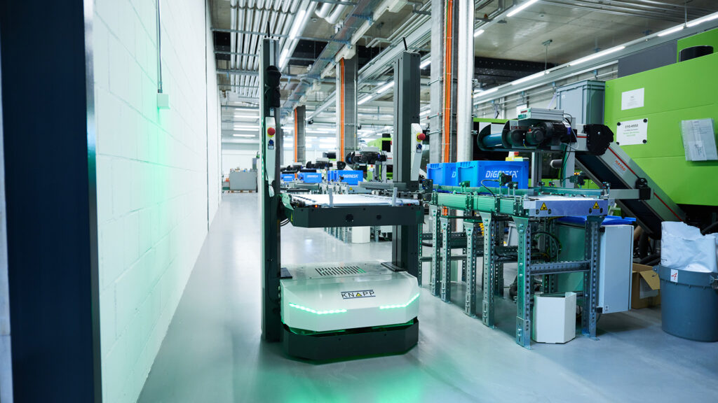 Un AGV RMA transporta materiales de forma autónoma a través de una instalación industrial limpia y moderna repleta de maquinaria, estanterías e iluminación superior.