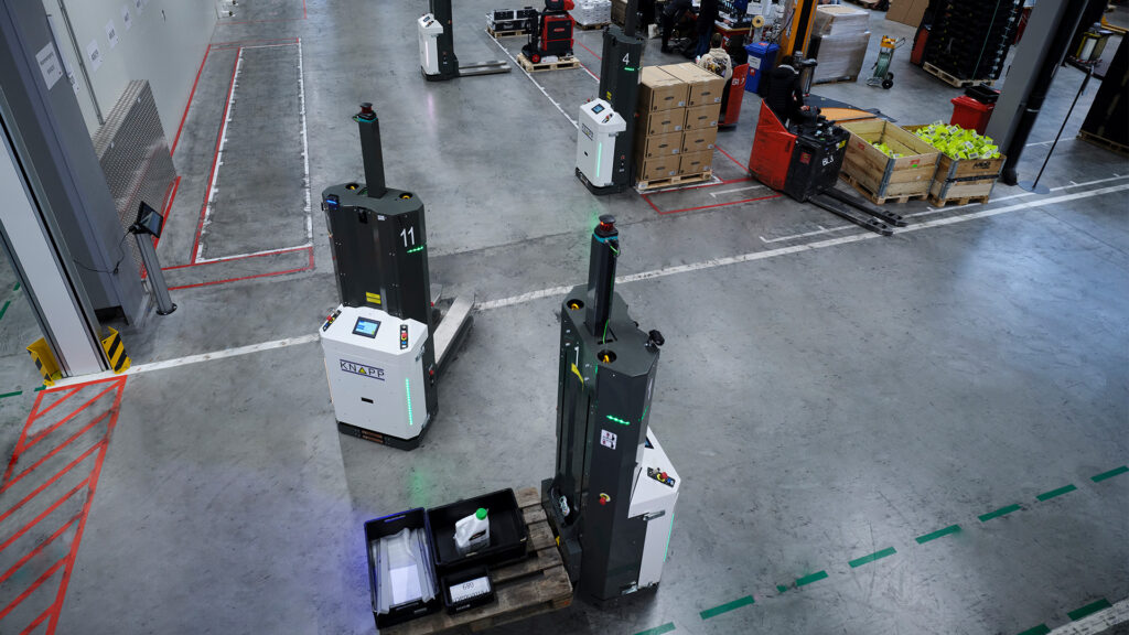 AGV AMR-Roboter transportieren Waren und Paletten in einem geräumigen Lager mit organisierten Lagerbereichen und sichtbaren Sicherheitsmarkierungen auf dem Boden.