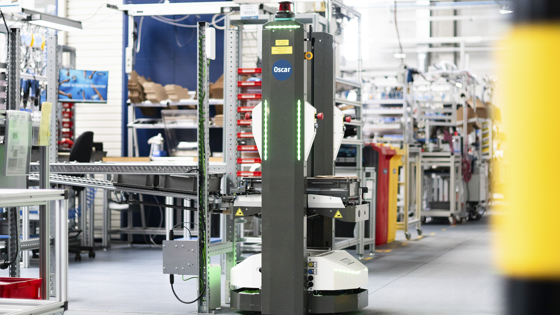 Ein autonomer mobiler Roboter namens Oscar befördert Güter in einem modernen Industrielager mit Regalen, Förderbändern und Maschinen.