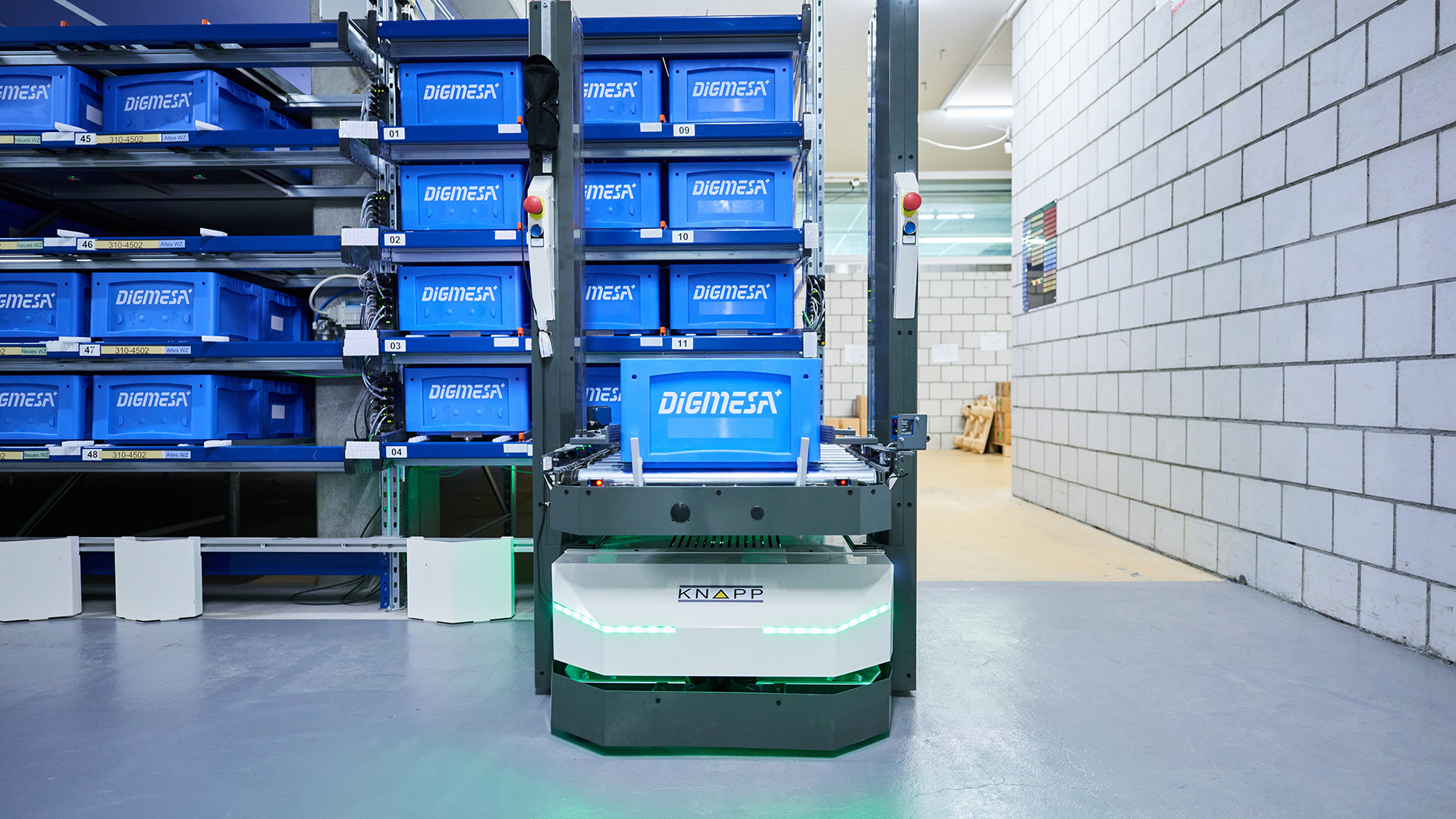 Un robot déplace des bacs DIGIMAX bleus dans un entrepôt où se trouvent des rayonnages à gauche et des murs en briques blanches à droite.