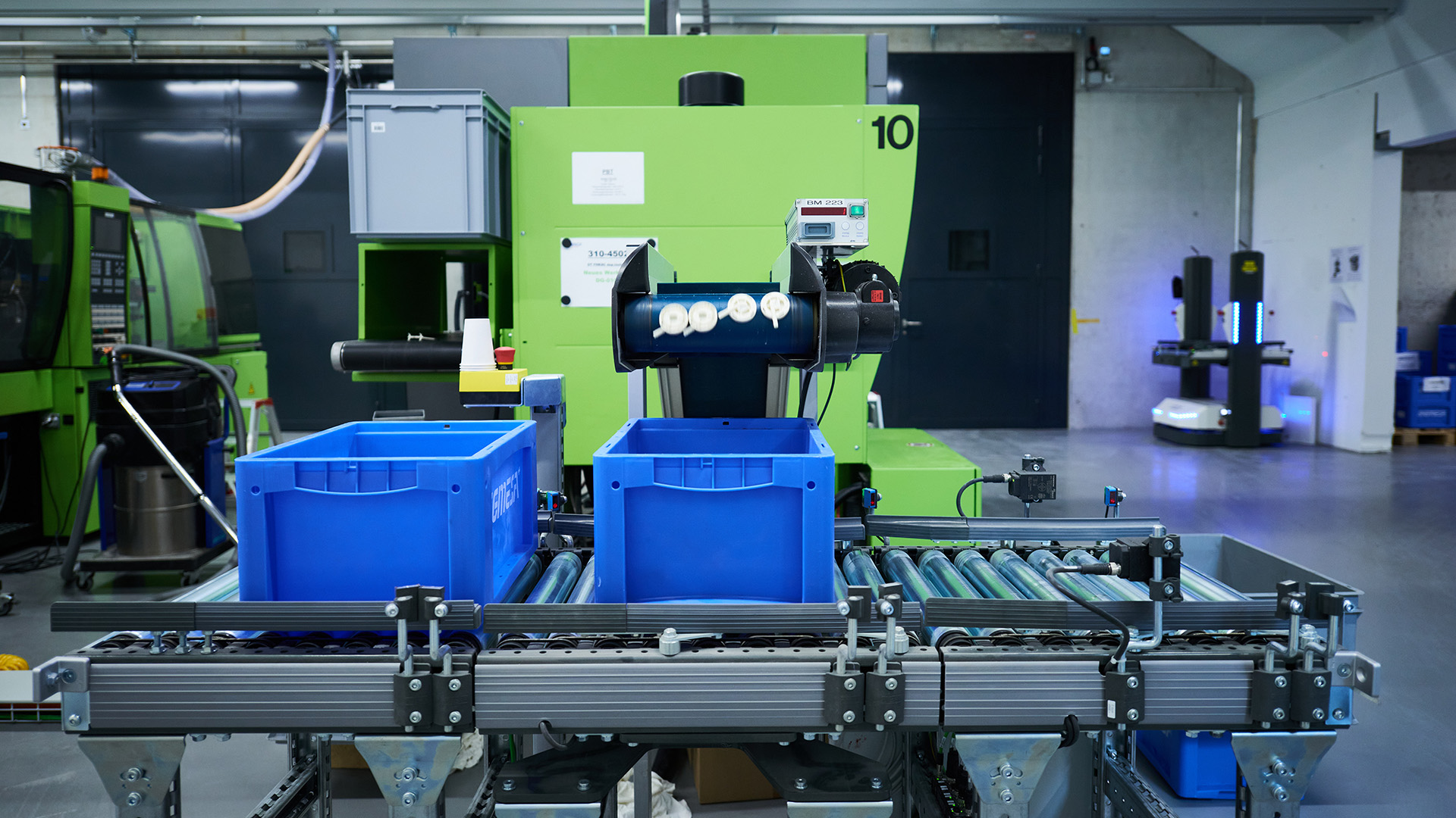 Des bacs en plastique bleus sur un tapis roulant, dans une usine industrielle moderne, avec des machines et des équipements verts en arrière-plan.