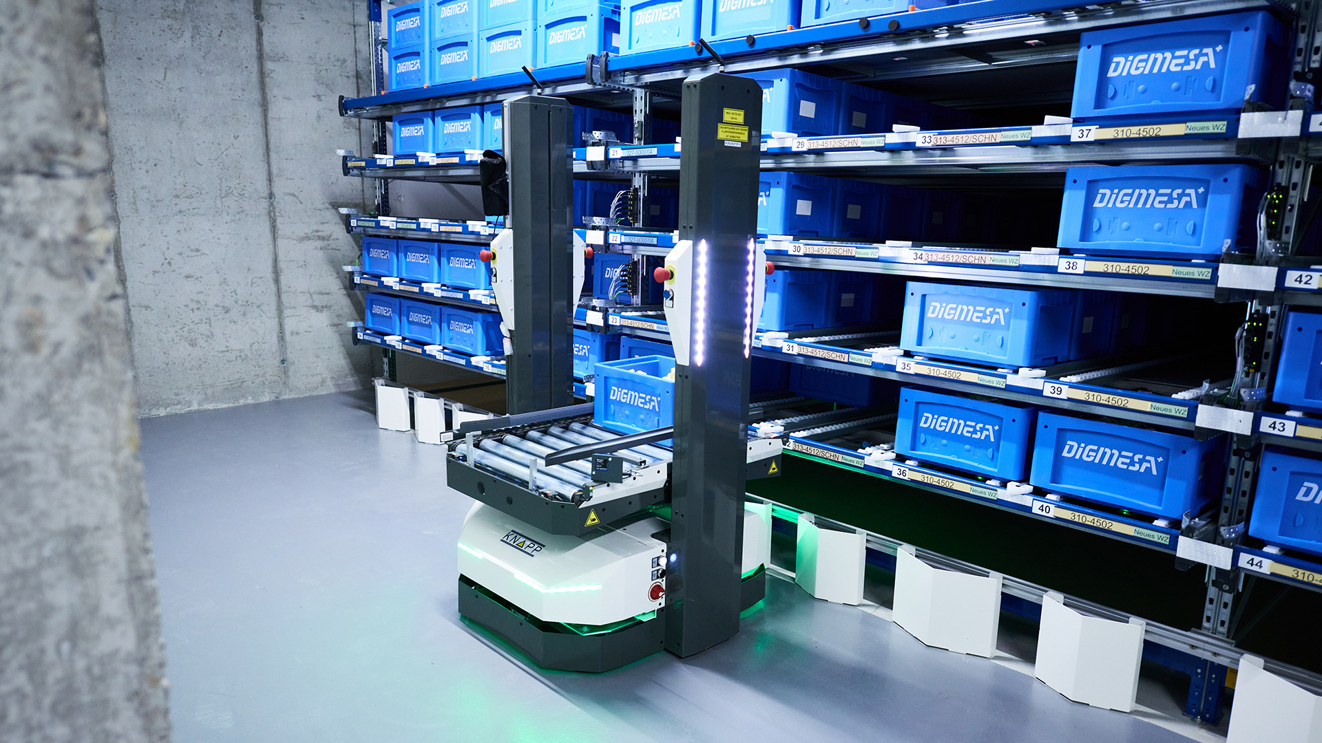 Dans un environnement industriel, un robot d'entrepôt équipé de rouleaux se trouve entre des rayonnages remplis de bacs de stockage bleus portant l'étiquette « DIGIMESA ».