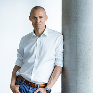 A man wearing a white shirt and jeans is leaning against a concrete column and smiling into the camera.