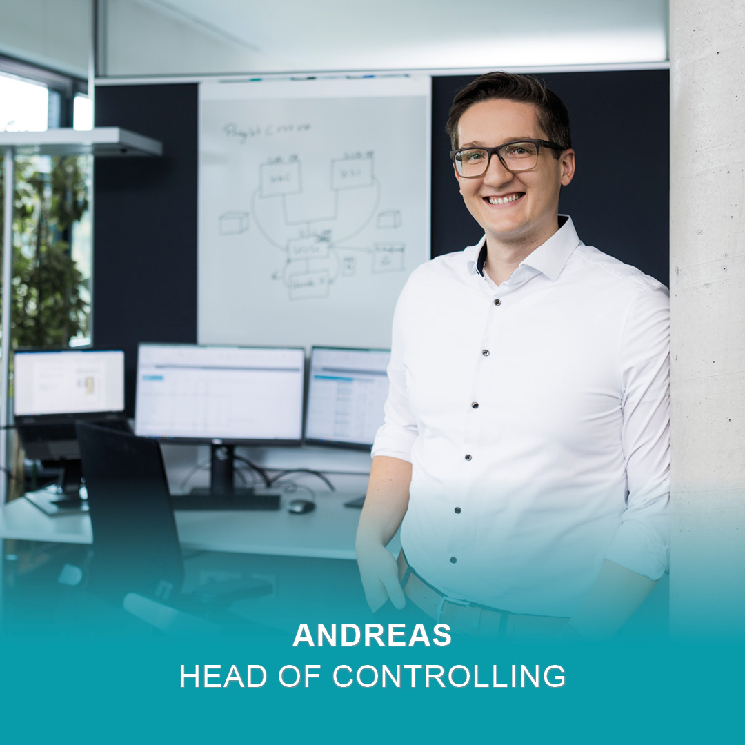 Ein lächelnder Mann mit Brille und weißem Hemd steht in einem modernen Büro für Controlling, umgeben von Computermonitoren und einem Whiteboard. Der Text auf dem Bild lautet: "ANDREAS HEAD OF CONTROLLING".