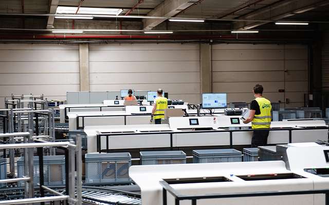 Des travailleurs portant des gilets de sécurité jaunes utilisent des ordinateurs et des équipements dans un grand entrepôt moderne ou un centre de distribution avec de hauts plafonds et un éclairage industriel.