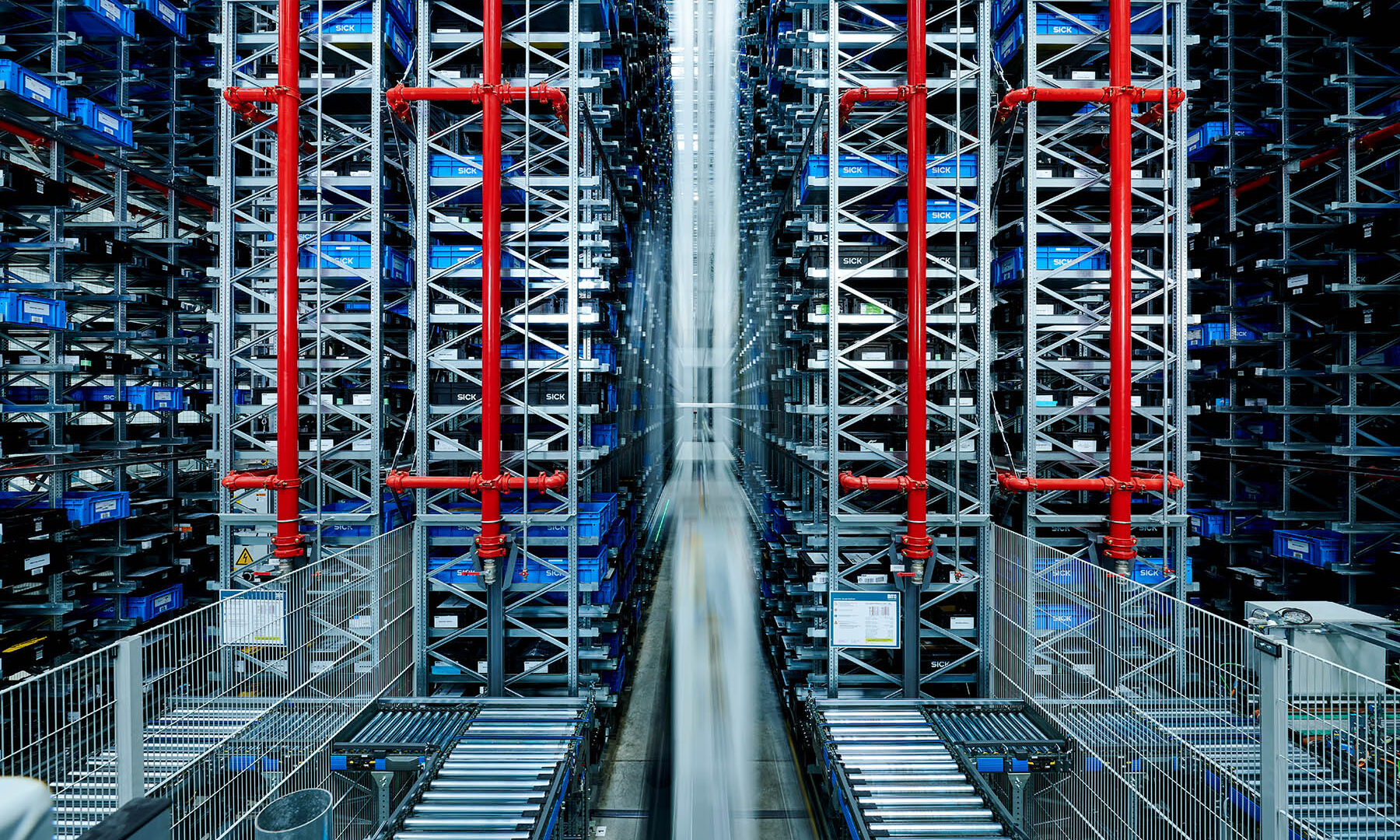 Vue d'un entrepôt automatisé avec de hautes étagères remplies de bacs bleus, une structure métallique, des tuyaux rouges et des bandes transporteuses en bas, créant une scène industrielle symétrique.