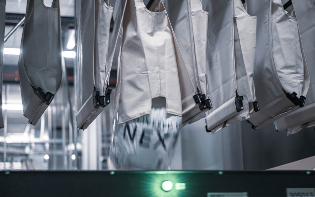 autopocket_inditex_tempe_2 Mehrere weiße Stoffsäcke hängen an einem automatisierten Sortiersystem in einer industriellen Umgebung. Eine AutoPocket in Bewegung enthält ein Paket mit der Aufschrift "NEXT".