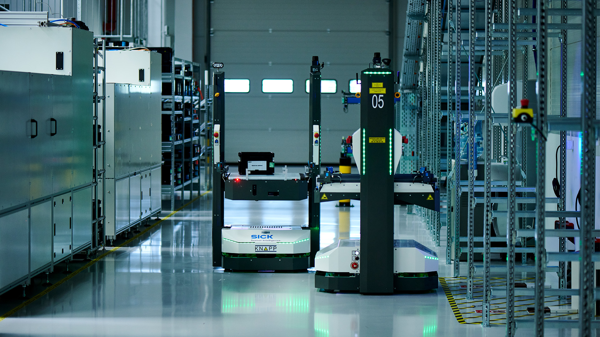 Deux robots autonomes naviguent dans une allée d'entrepôt moderne et bien éclairée, bordée d'étagères et d'équipements industriels.