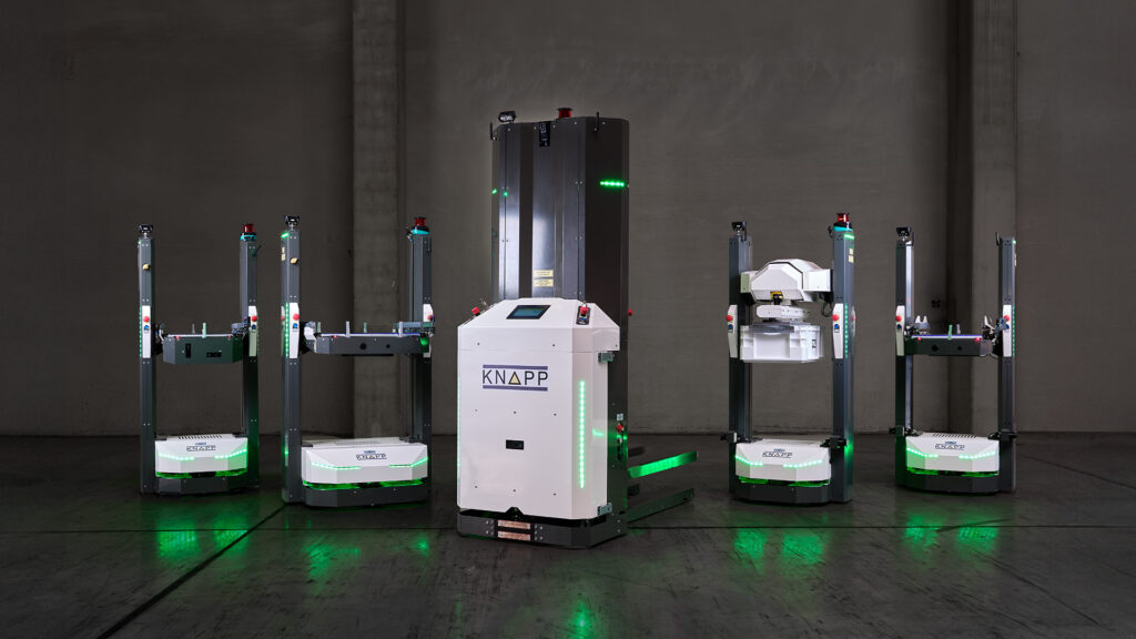 Dans un espace industriel peu éclairé, cinq robots d'entrepôt automatisés dotés de lumières vertes sont alignés à côté d'un grand robot blanc portant l'inscription "KNAPP".