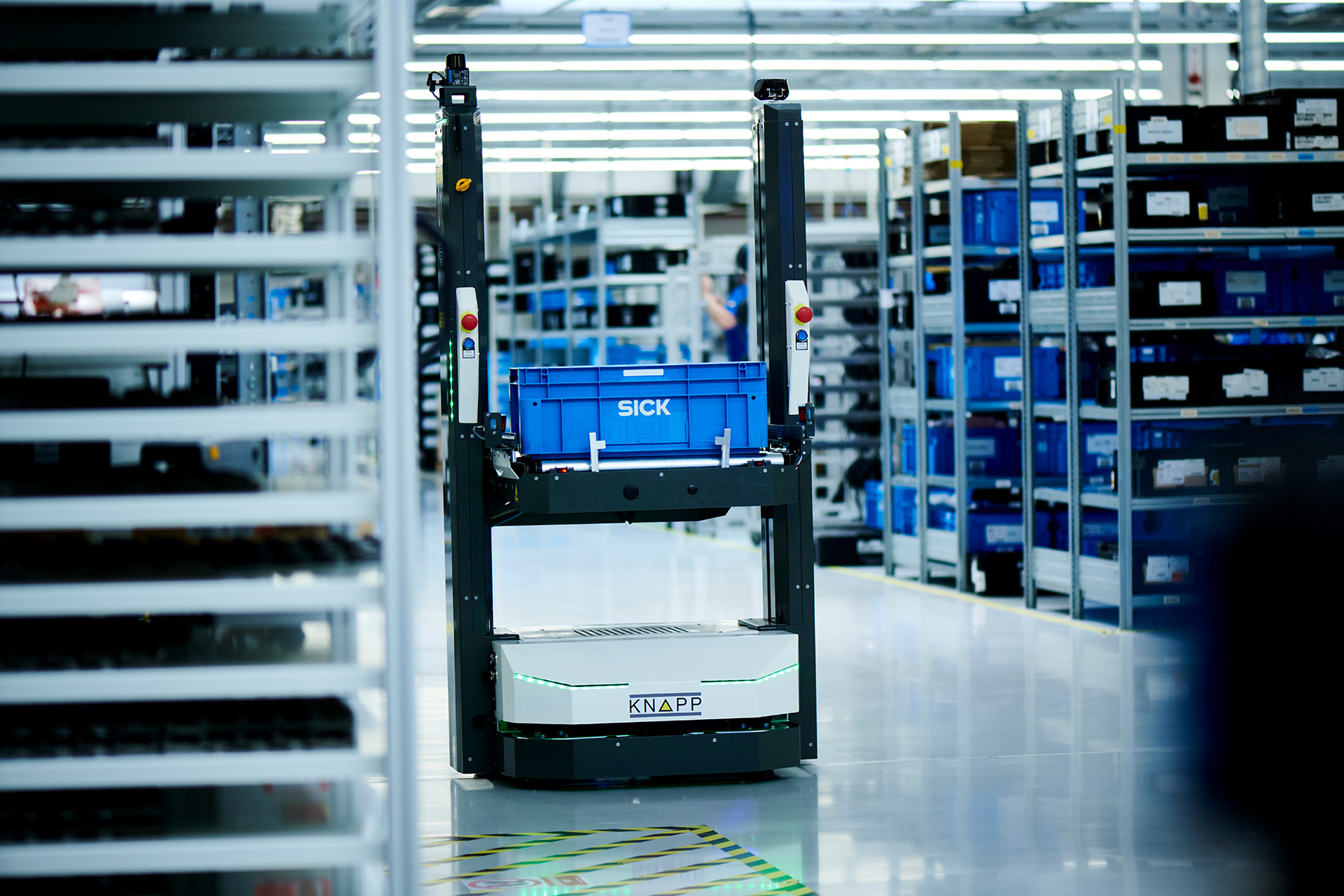 Un chariot robotisé transporte un conteneur bleu dans un entrepôt moderne avec des étagères de pièces et un éclairage zénithal lumineux.