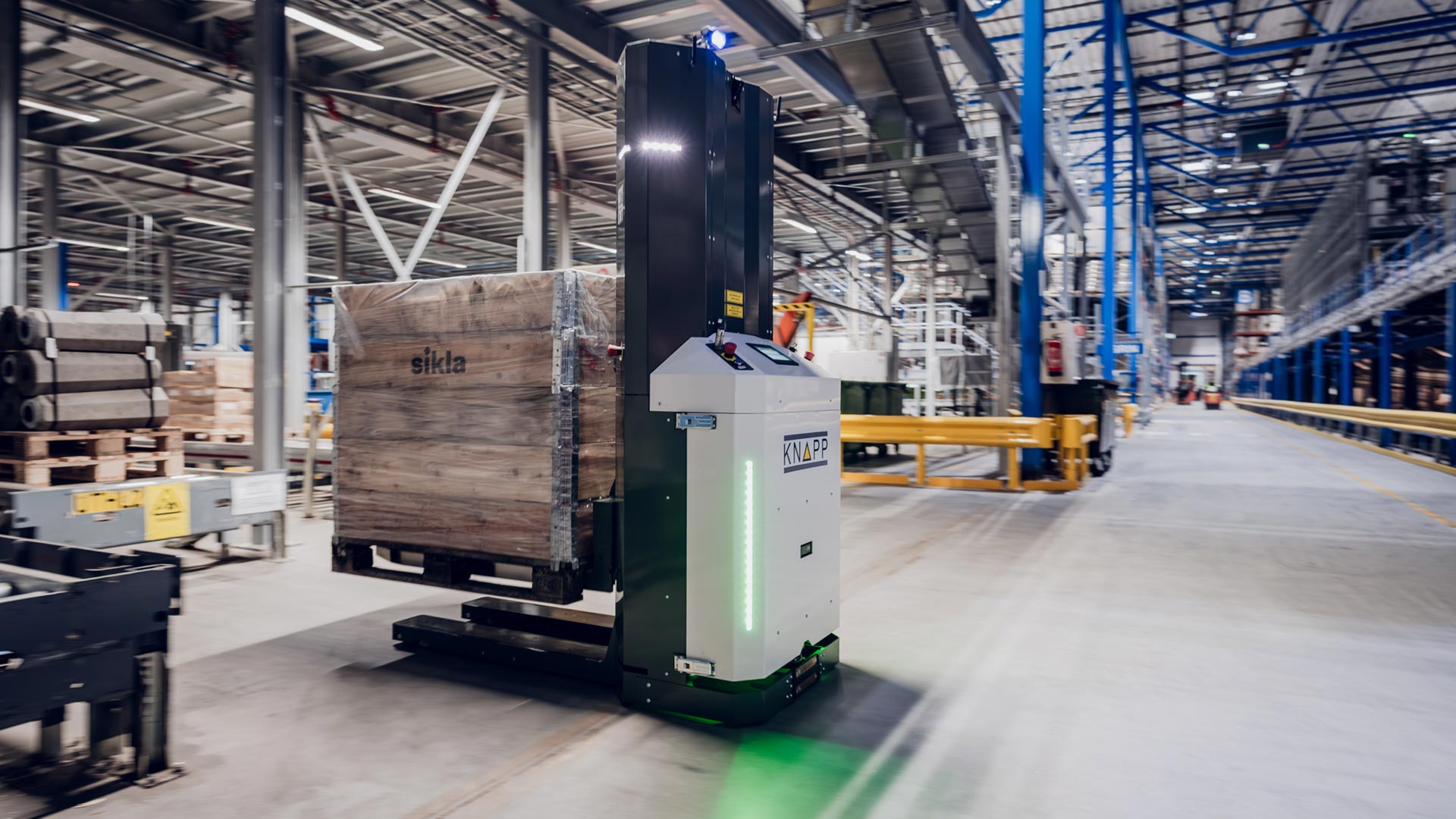 Brodrene-Dahl_Web_fork_2 Un vehículo de guiado automático transporta un palé de madera en un gran almacén moderno con estanterías y equipos industriales.