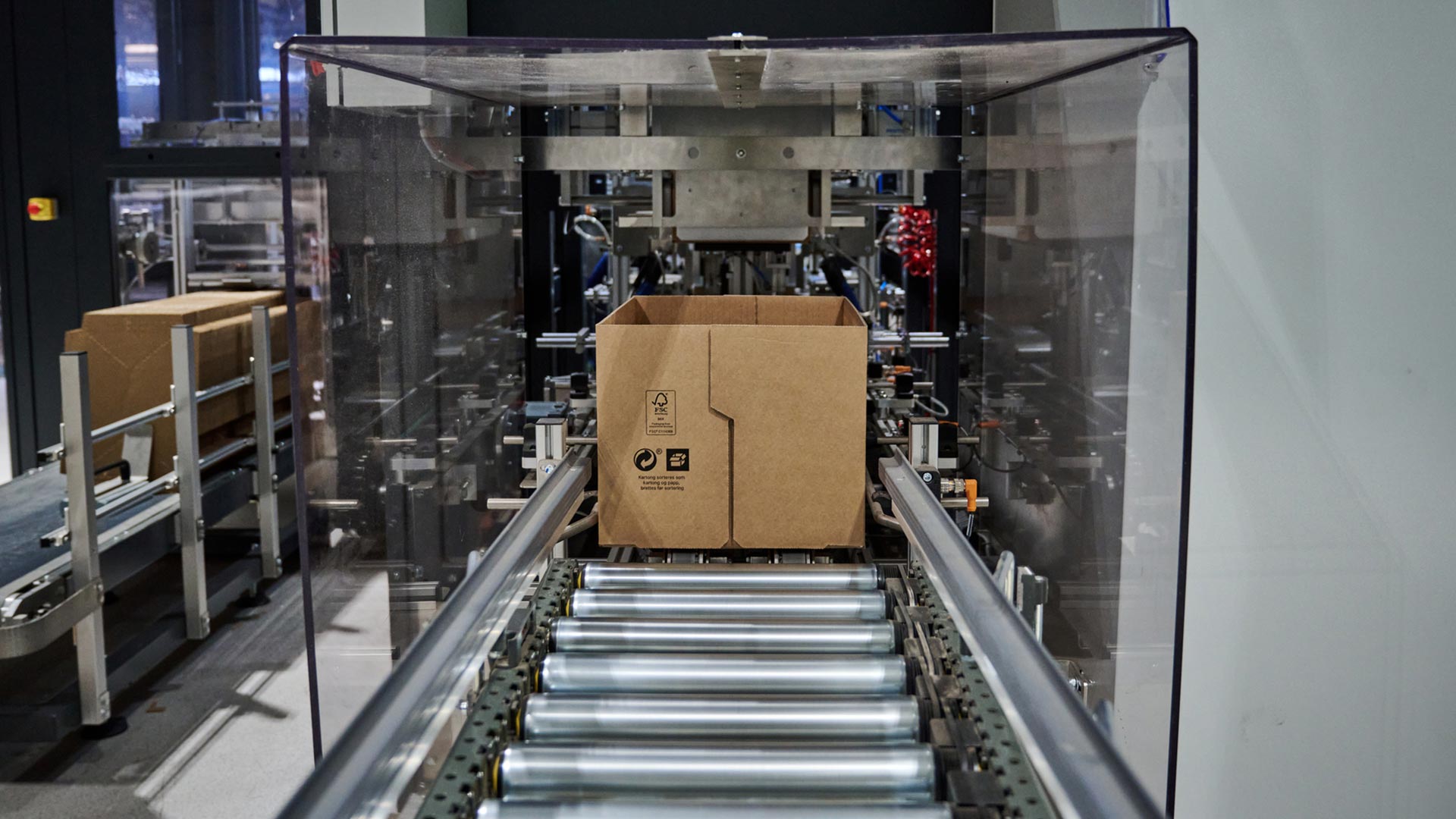 Brodrene-Dahl_Web_Lagerprozesse_3 Una caja de cartón se desplaza por una cinta transportadora automatizada dentro de una instalación de envasado o fabricación.
