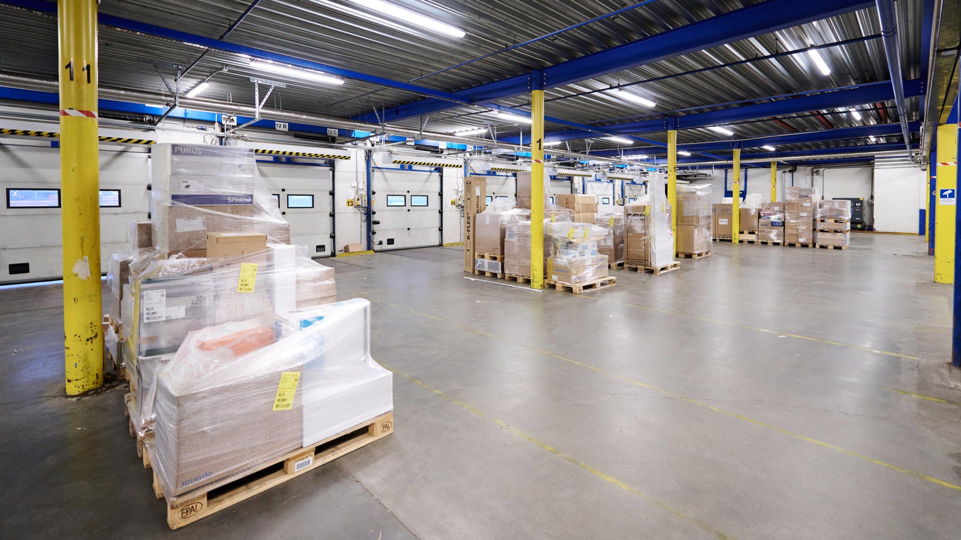 Brodrene-Dahl_Web_Lagerprozesse_4 Interior de almacén con varios palés de mercancías en cajas envueltas en plástico, columnas de soporte amarillas y puertas de muelle de carga al fondo.