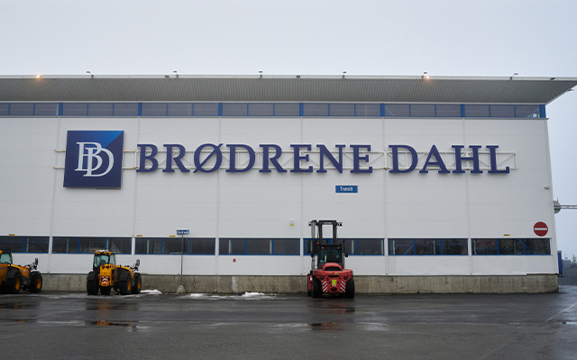 A large industrial building with the sign "BRØDRENE DAHL" and logo on the front. Several construction vehicles are parked outside the building on a cloudy day.